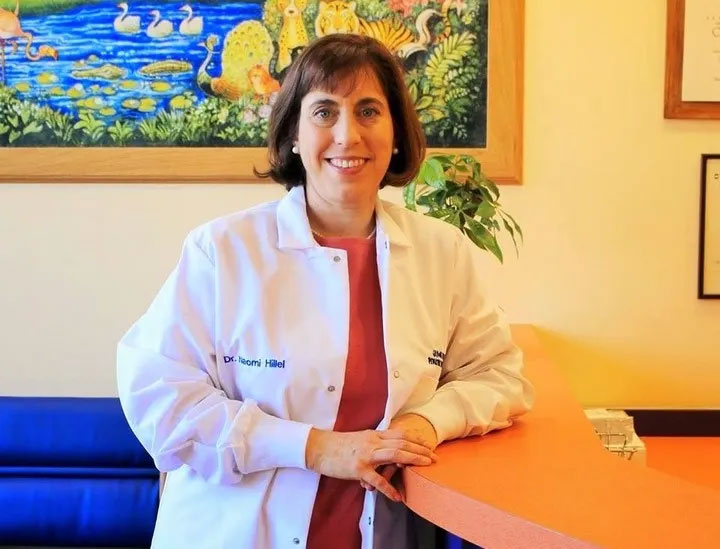 The image shows a woman standing behind a desk with a nameplate, wearing a white lab coat over a red shirt, posing for a photograph.